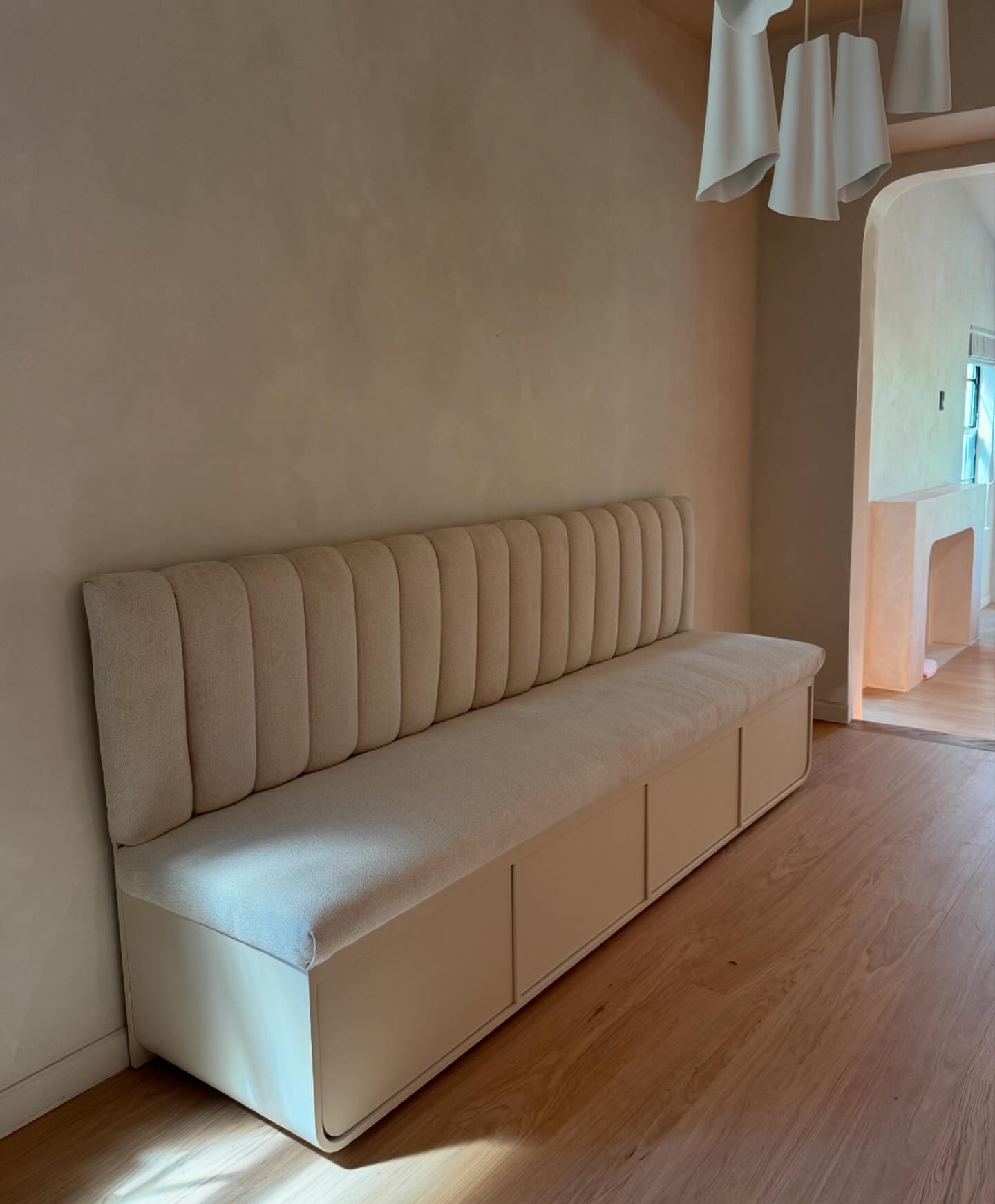 A handcrafted channel-back banquette in pale linen, with a white cabinet base, in a sunlit room.