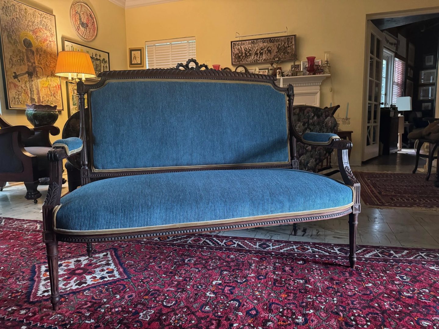 A carved dark-wood settee with a ribbed blue velvet seat and back, on a red patterned rug in a yellow living room.
