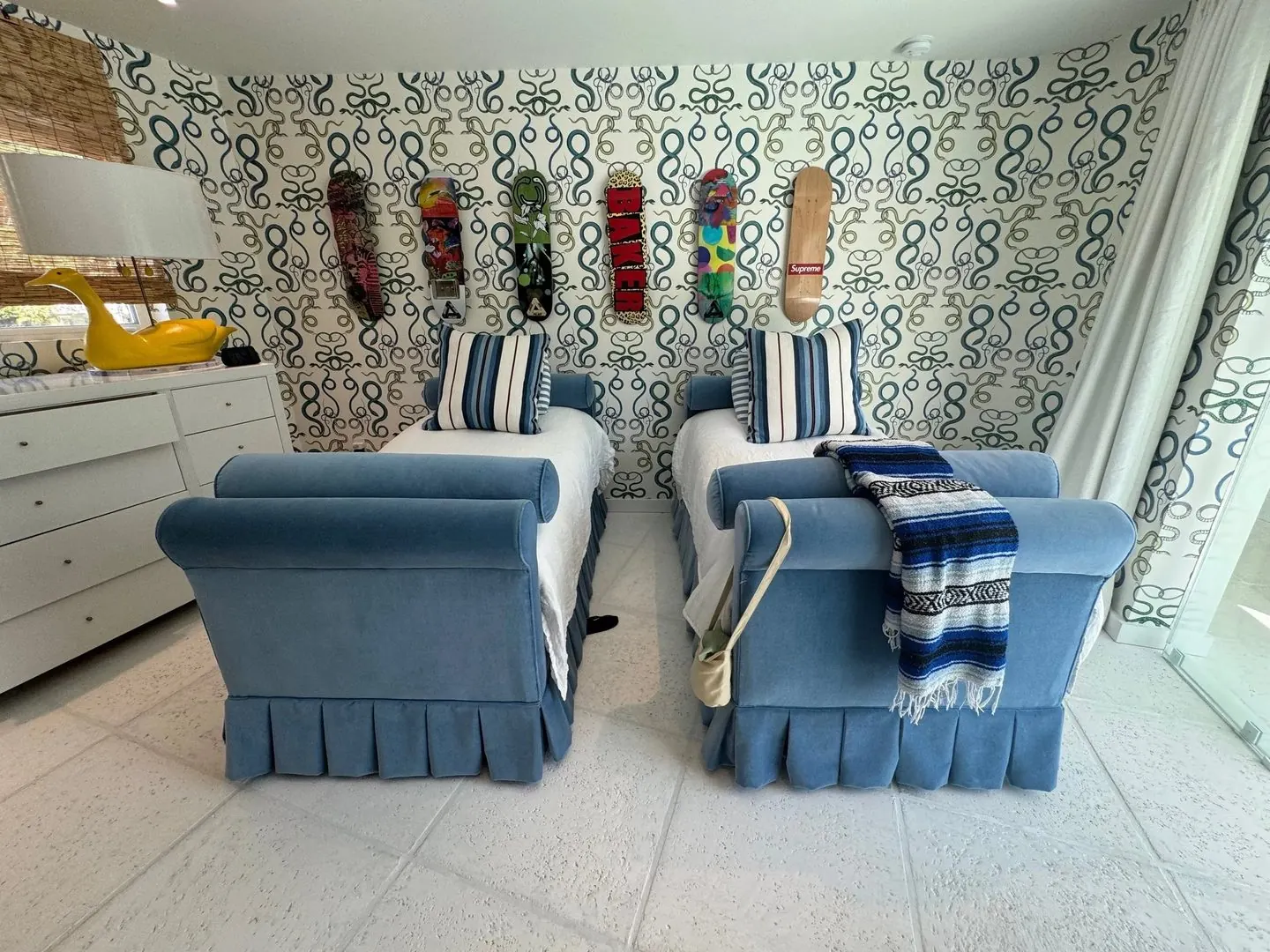 Two twin beds with rolled blue velvet frames and pleated skirts, with blue-and-white striped pillows, against green-and-white patterned wallpaper hung with skateboards.