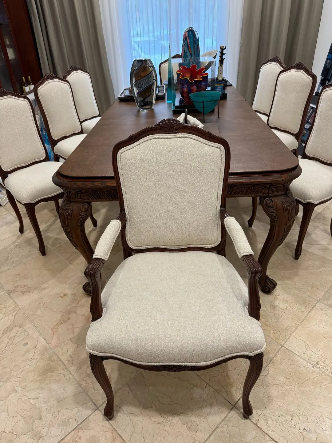 Carved wood dining chairs, including an armchair at the head, covered in cream fabric around a dark wood table.