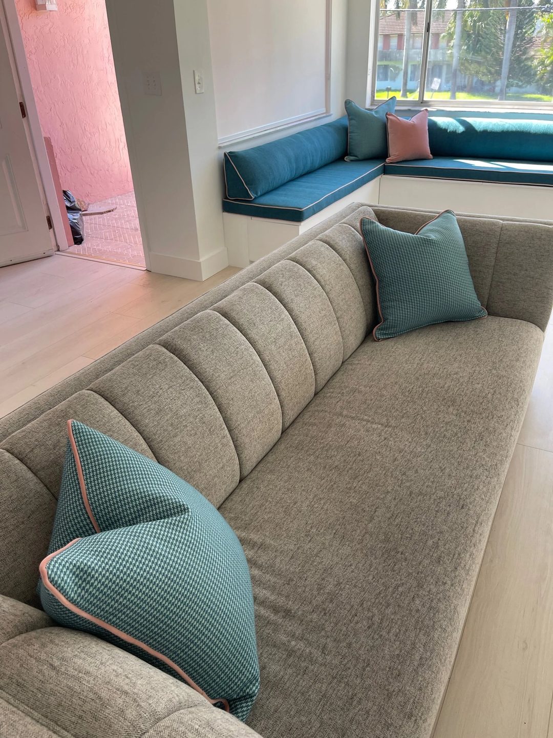 A long grey channel-back sofa with teal houndstooth pillows piped in pink, and a teal built-in window seat behind it.