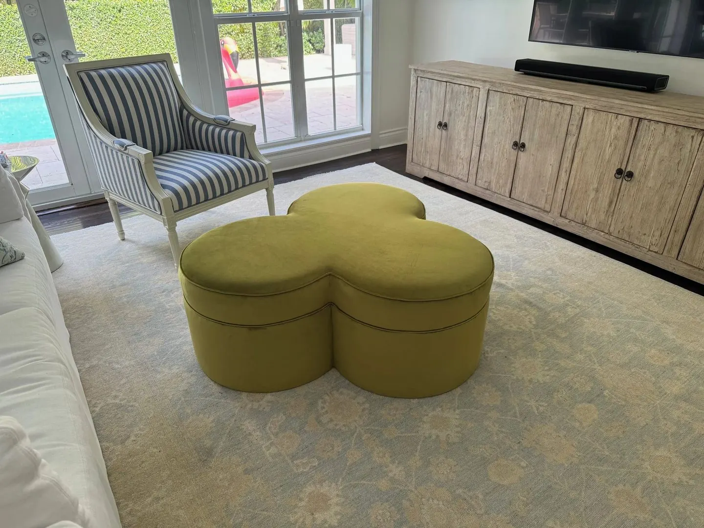 A clover-shaped ottoman in chartreuse velvet on a pale rug, with a blue-and-white striped armchair behind it.