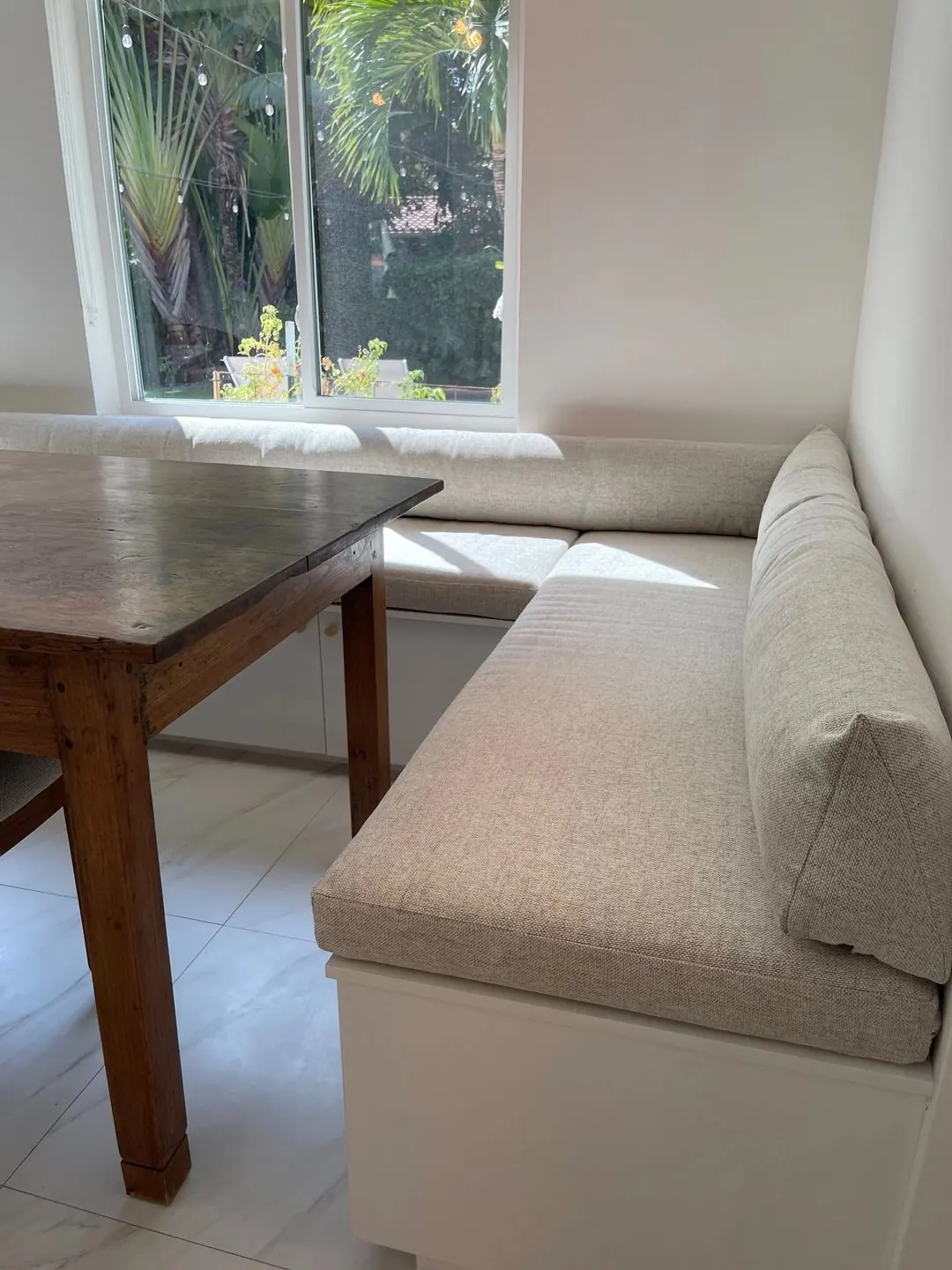 An L-shaped built-in banquette with oatmeal seat and back cushions on a white base, beside a wooden dining table and a window.