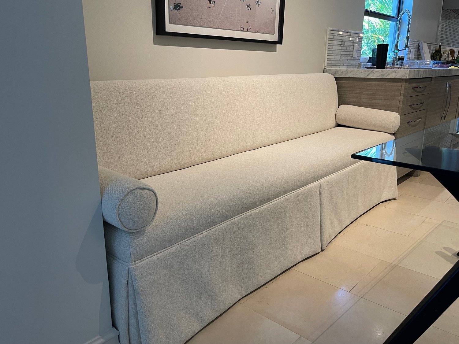 A long skirted banquette in off-white textured fabric with round bolsters at each end, beside a kitchen.