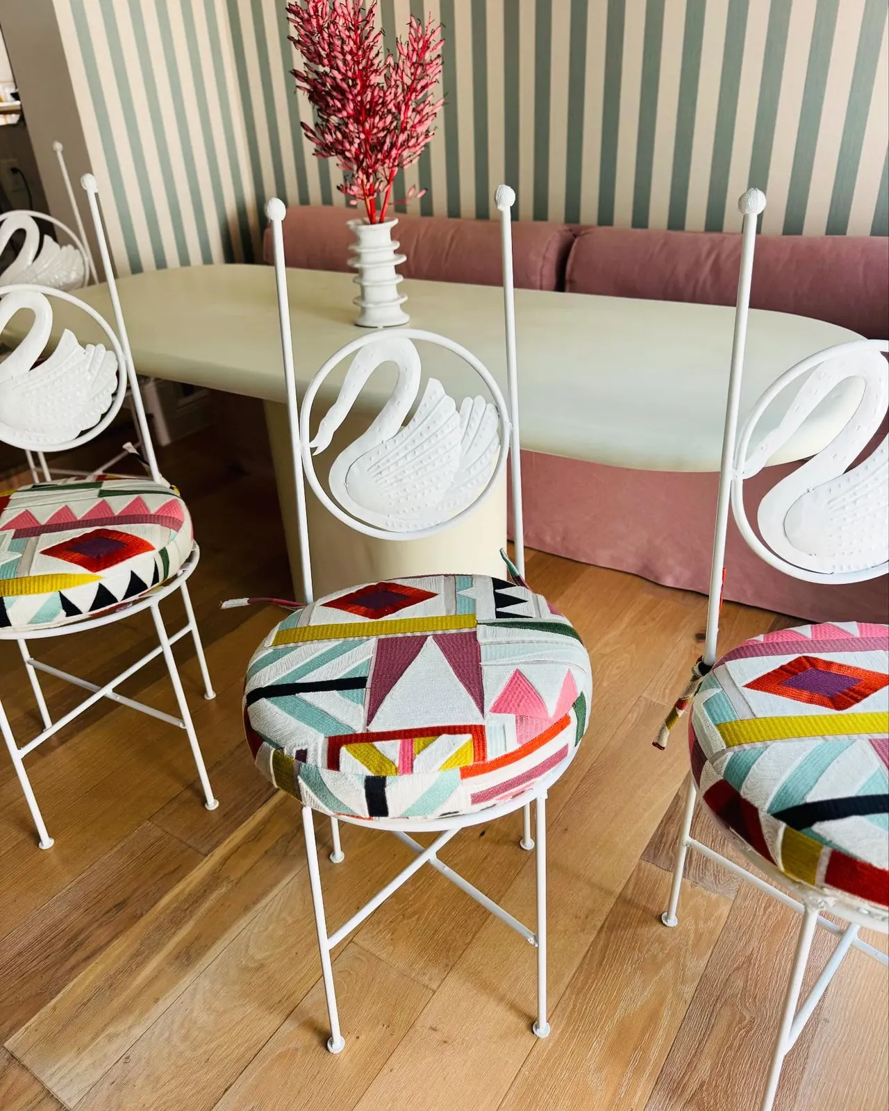 Four white swan-back chairs with bright geometric seat cushions around a pale dining table, with a dusty pink banquette behind them.