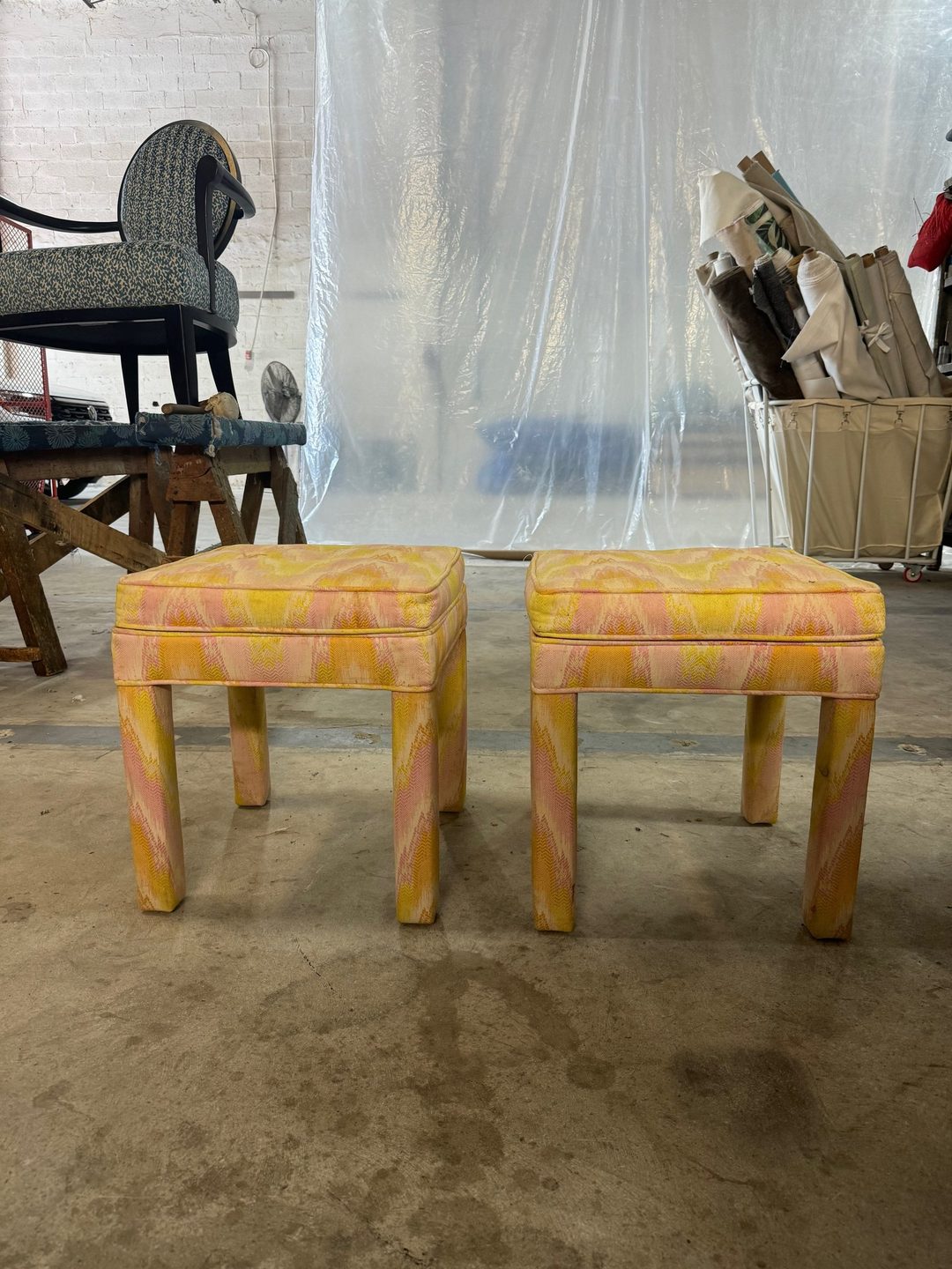 Two fully upholstered square stools in a pink and yellow flame-stitch fabric, standing on the workshop floor.