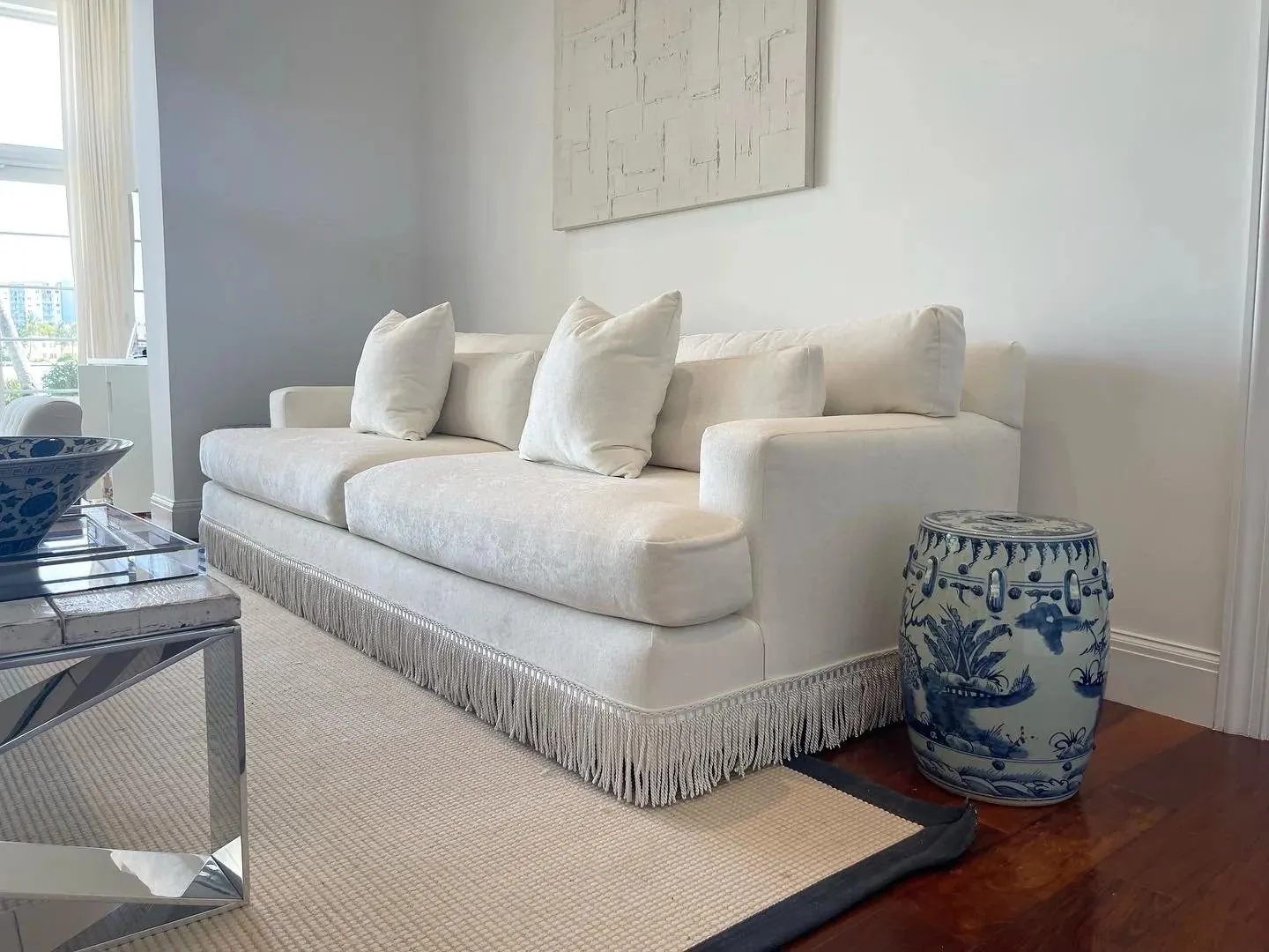 A white sofa with a long bullion fringe along its base, with a blue-and-white ceramic garden stool beside it.