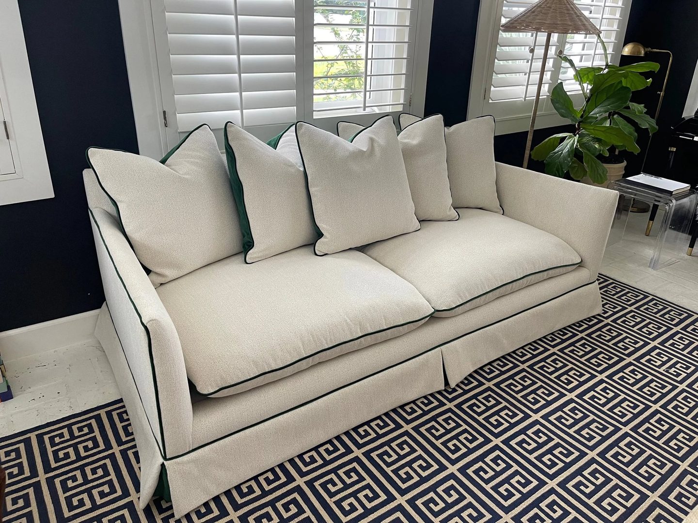 A skirted cream sofa with dark green piping and matching pillows, on a navy Greek-key rug.