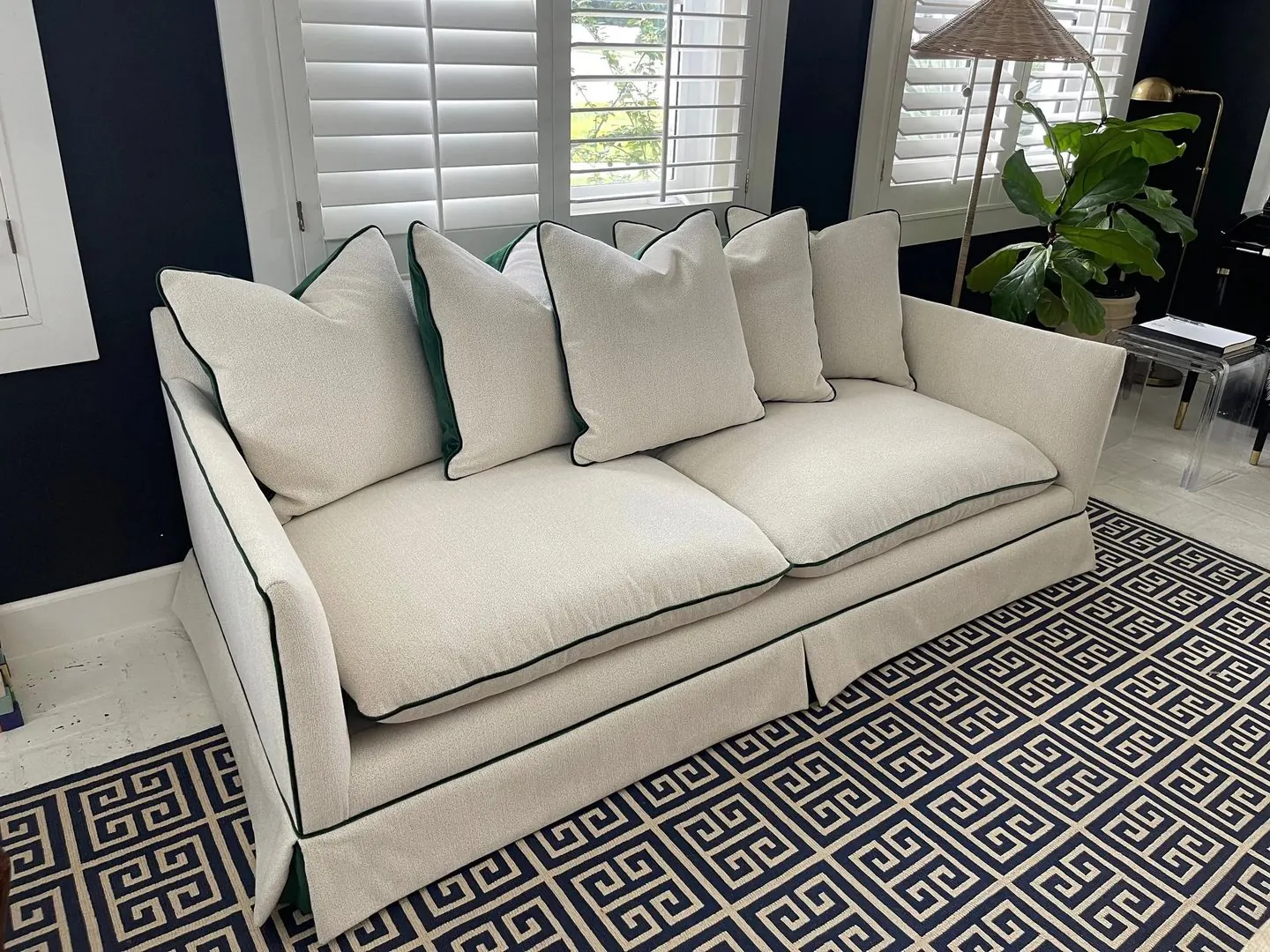 A skirted cream sofa with dark green piping and matching pillows, on a navy Greek-key rug.