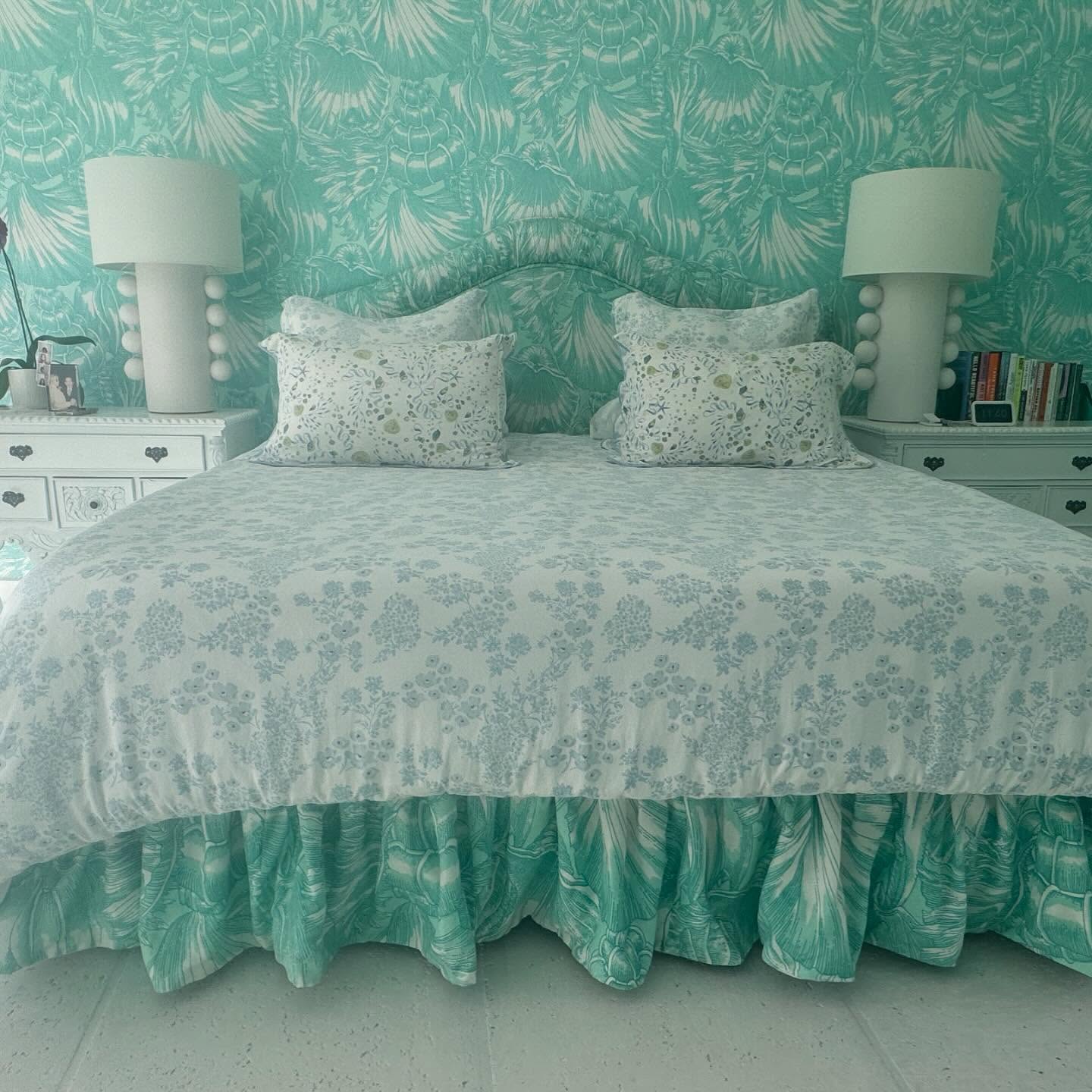A bed with a curved shirred headboard, floral pillows, and a turquoise ruffled bed skirt against matching wallpaper.
