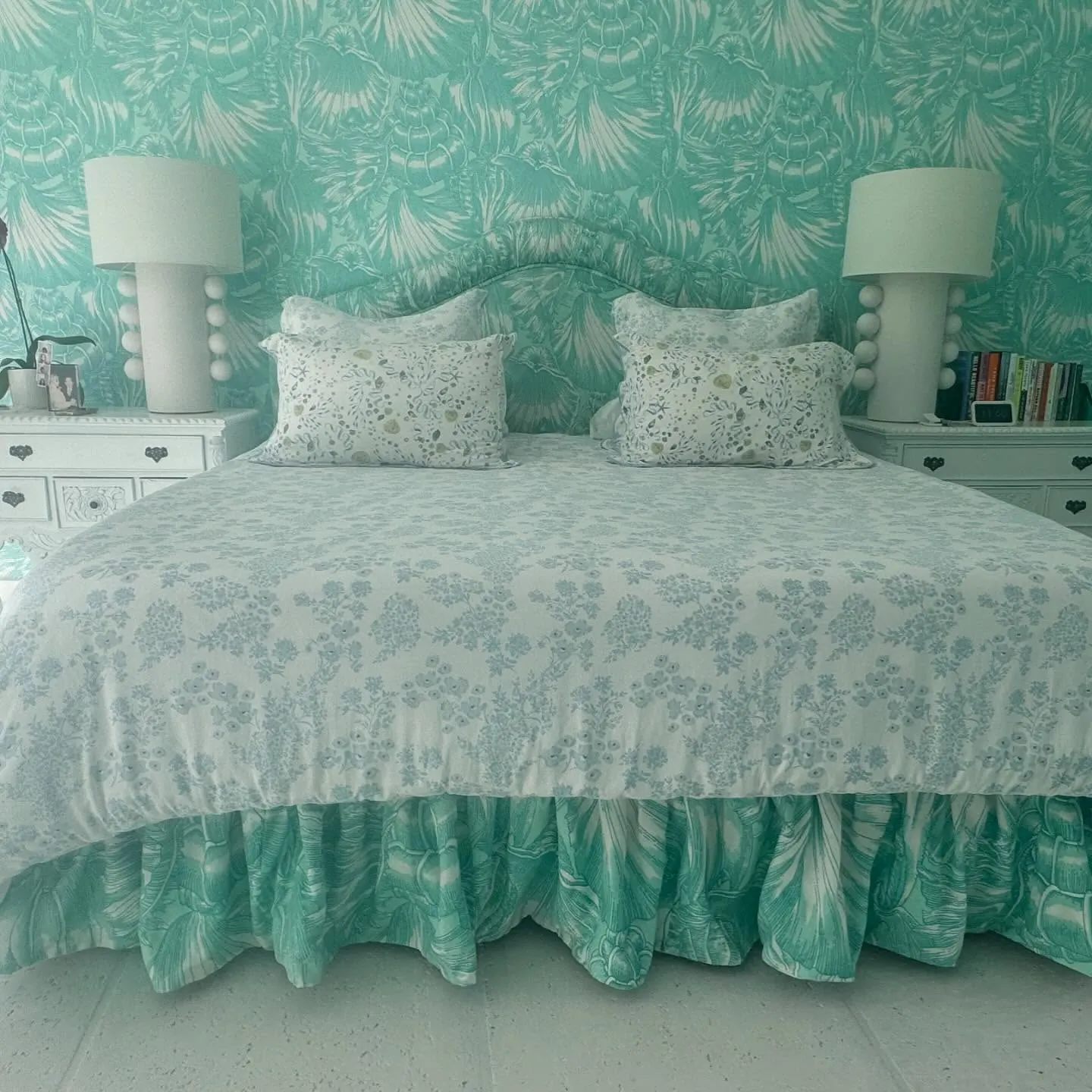 A bed with a curved shirred headboard, floral pillows, and a turquoise ruffled bed skirt against matching wallpaper.