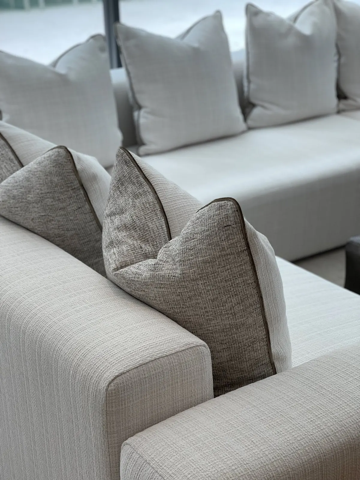 Close-up of a textured white sofa arm with grey tweed pillows piped in brown, and more white pillows along the sectional behind.