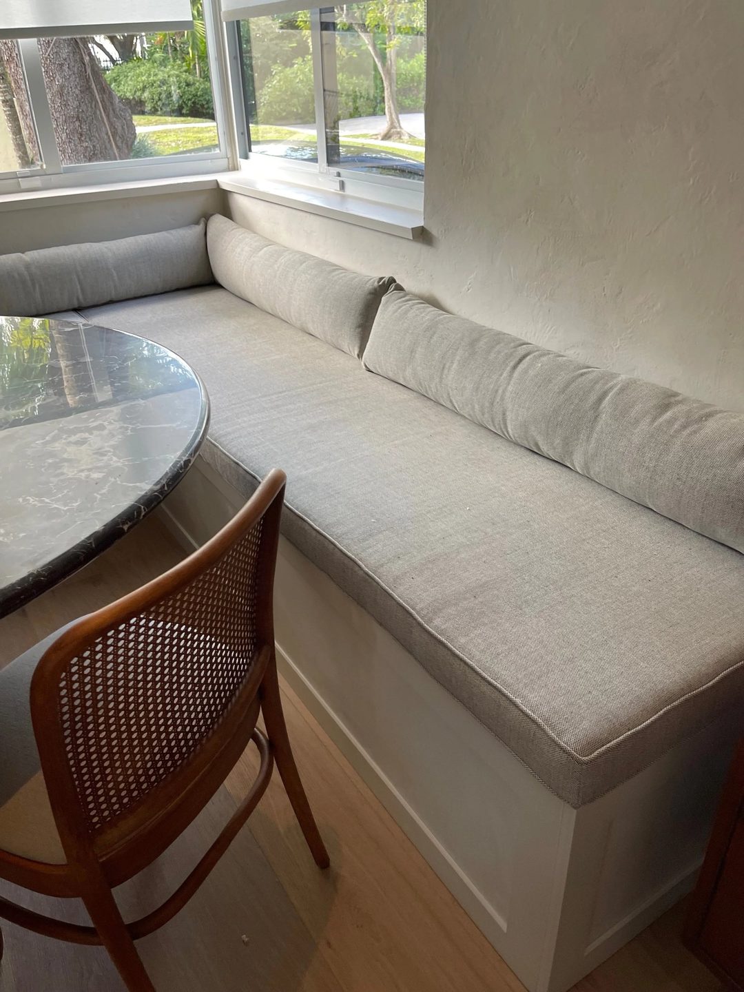 A built-in window seat with a grey linen cushion and long bolsters, beside a round marble table and a cane chair.