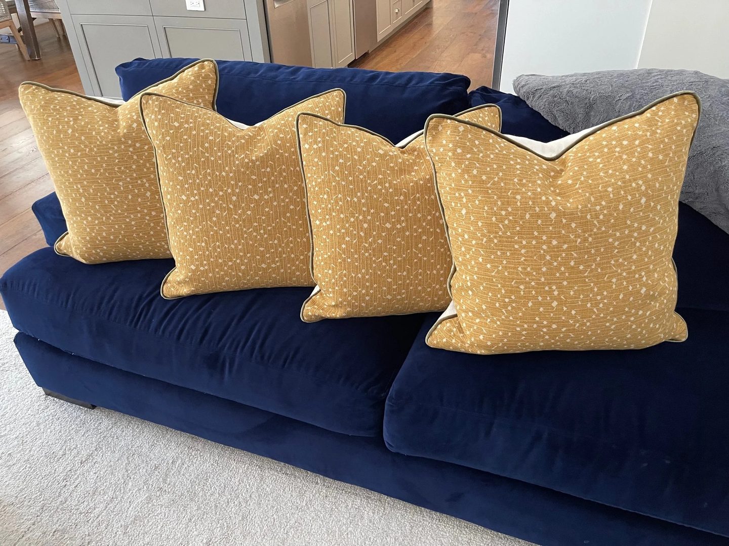 Four mustard pillows with a small white pattern and piped edges, lined up on a navy velvet sofa.