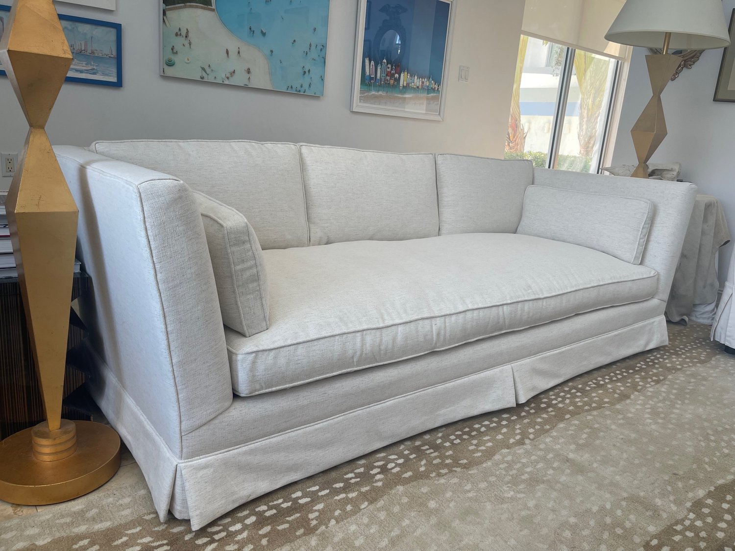 A tailored sofa in white textured fabric with a skirted base, on a spotted rug between two gold lamps.