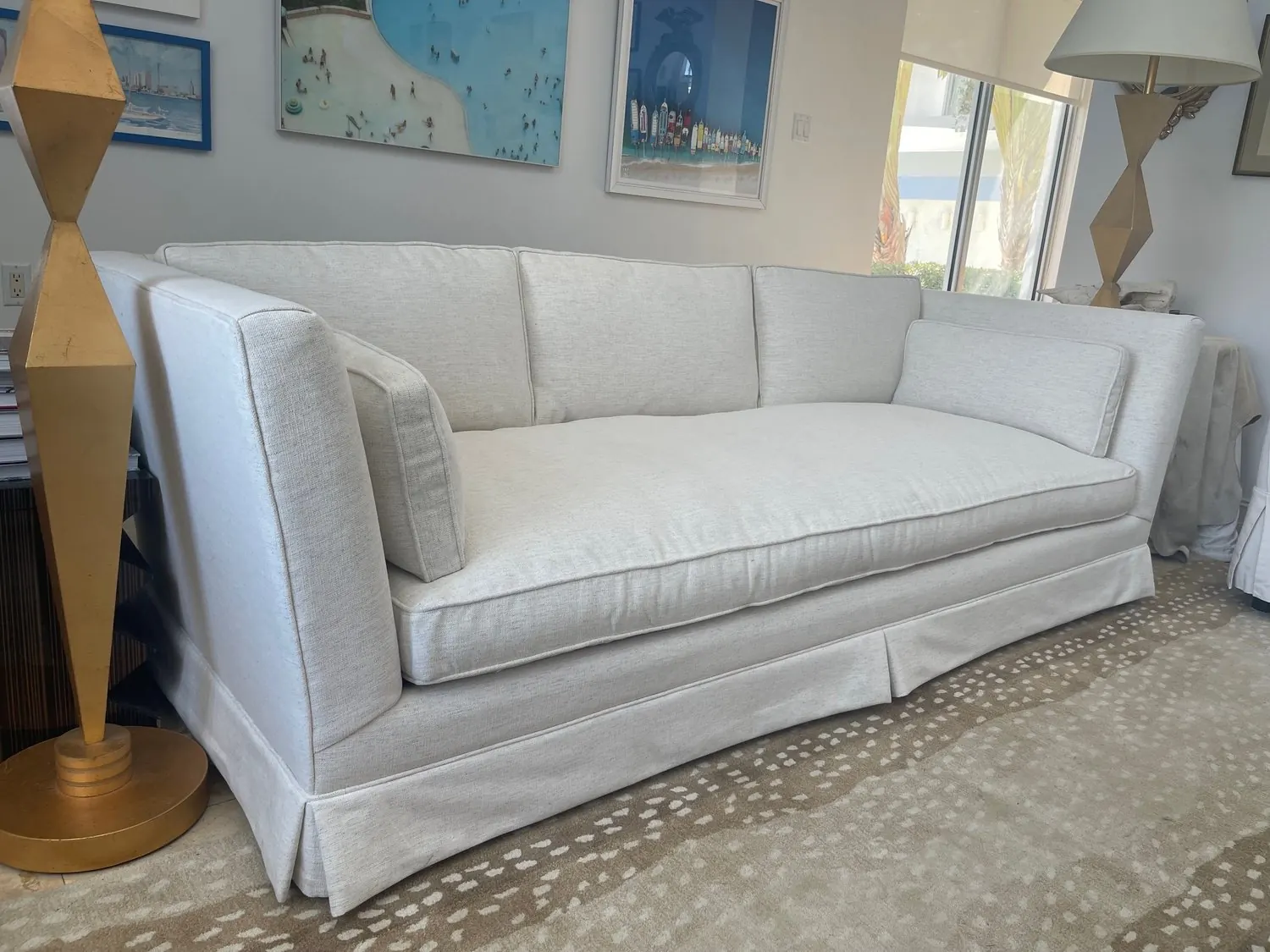 A tailored sofa in white textured fabric with a skirted base, on a spotted rug between two gold lamps.