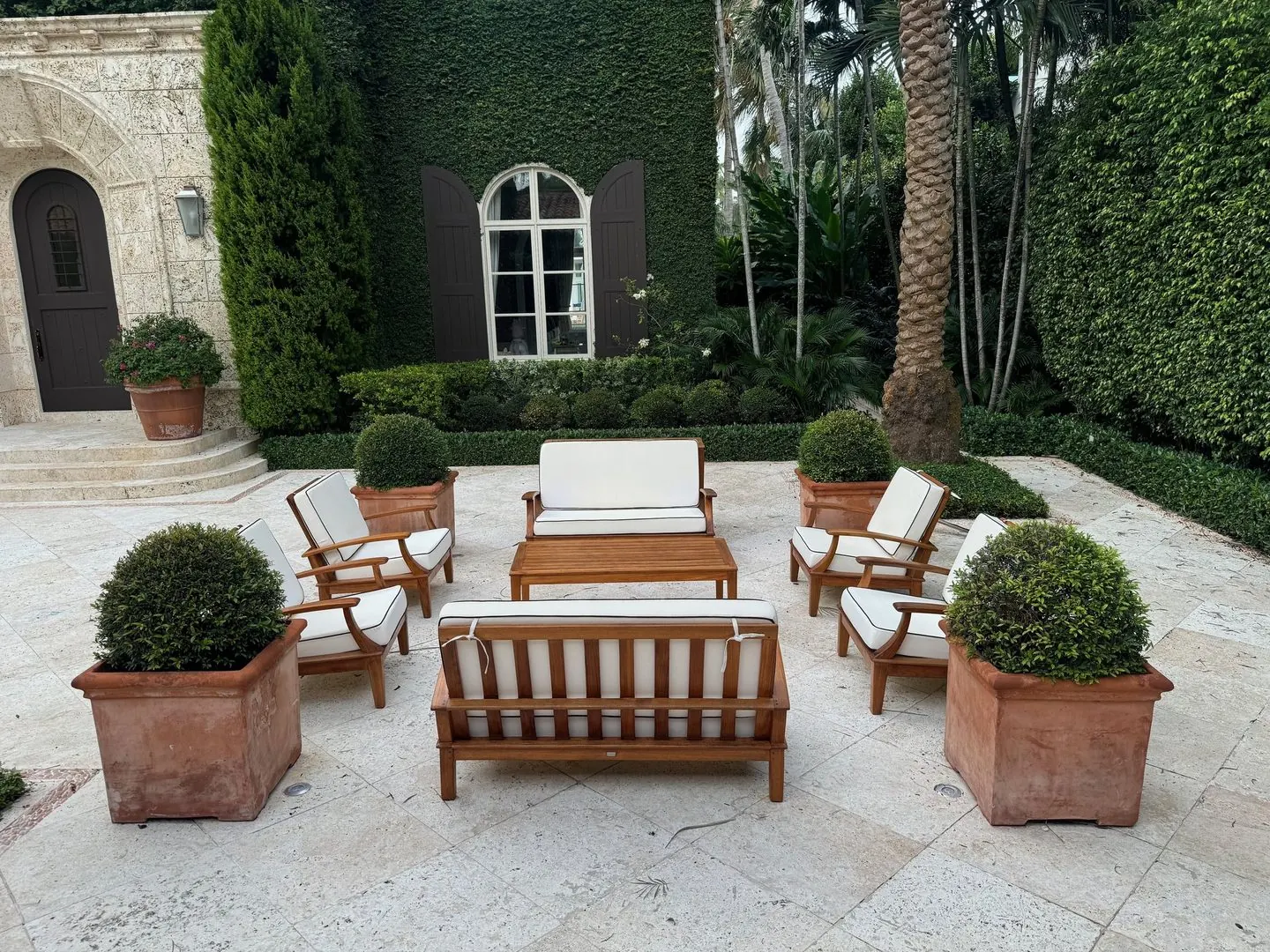 Teak outdoor sofas and armchairs with white cushions around a coffee table in a stone courtyard, with potted topiaries.