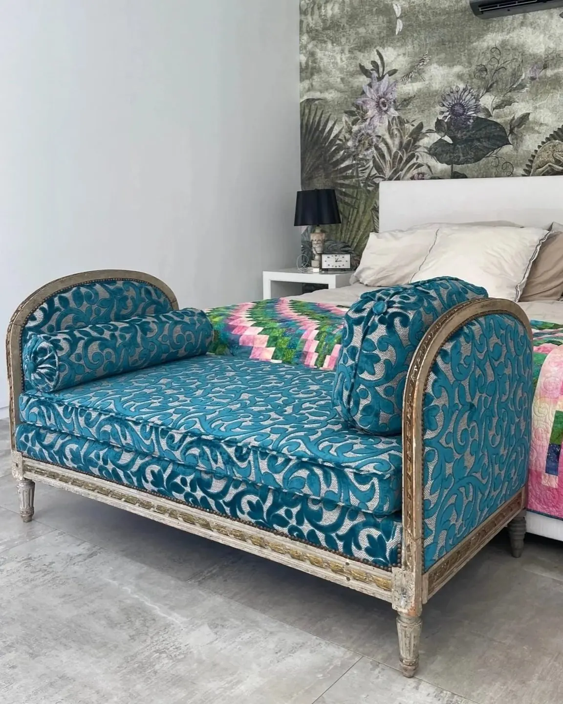 A painted wood daybed upholstered in teal and silver cut velvet, with a matching bolster and pillow, at the foot of a bed with a patchwork quilt.