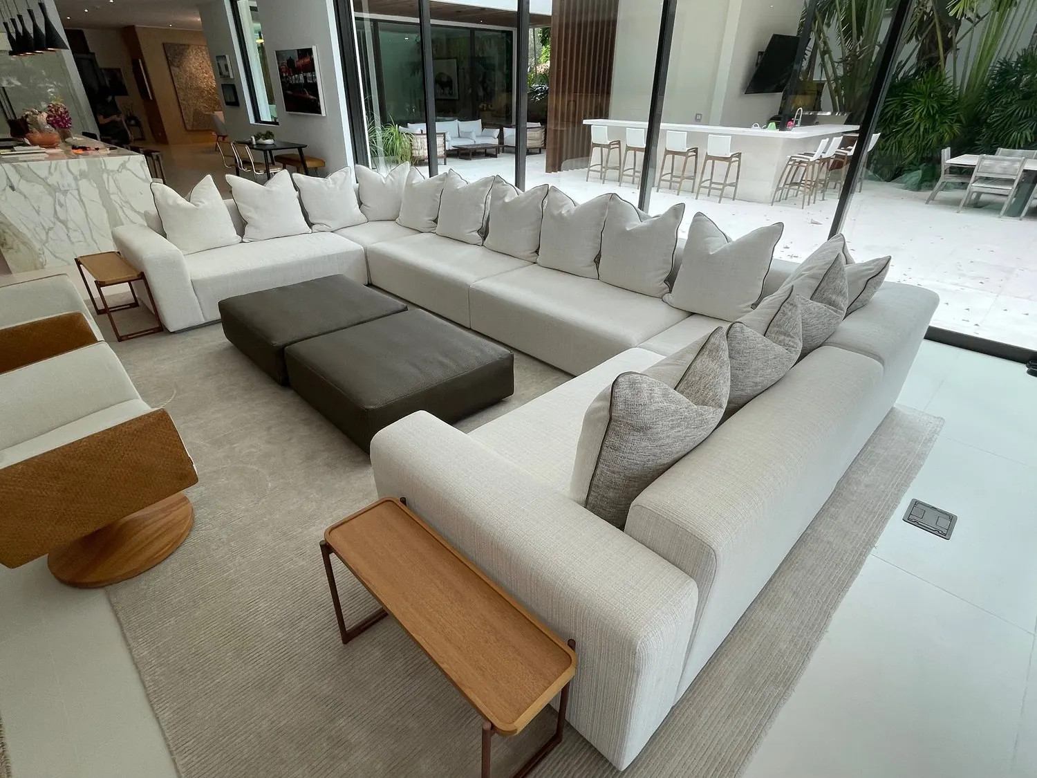 A large U-shaped white sectional with grey and white pillows around two charcoal ottomans, open to a pool terrace.