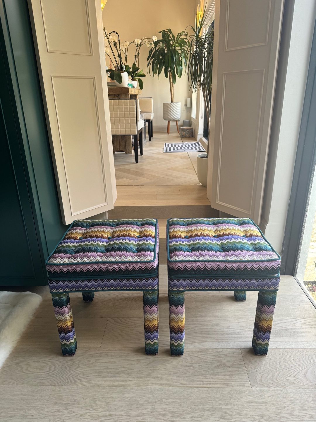 Two upholstered stools in a multicoloured zigzag knit, with tufted tops and teal piping, standing in a doorway.