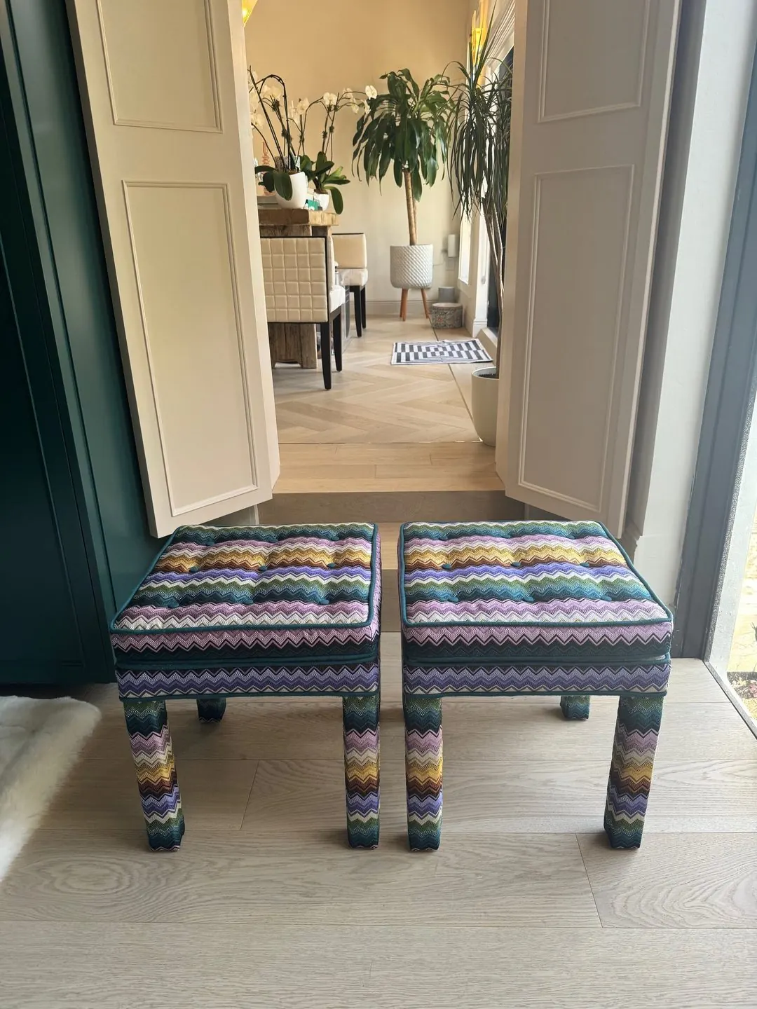 Two upholstered stools in a multicoloured zigzag knit, with tufted tops and teal piping, standing in a doorway.